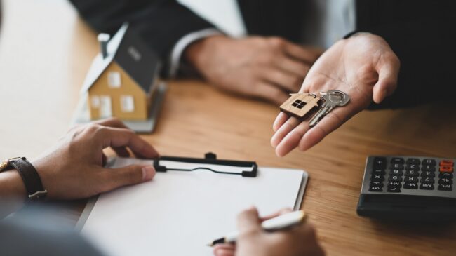 Cropped shot of Real estate agent giving a property key to his customer while signing an agreement on clipboard at the modern wooden table. Realtor/Broker/Seller/Dealer concept.